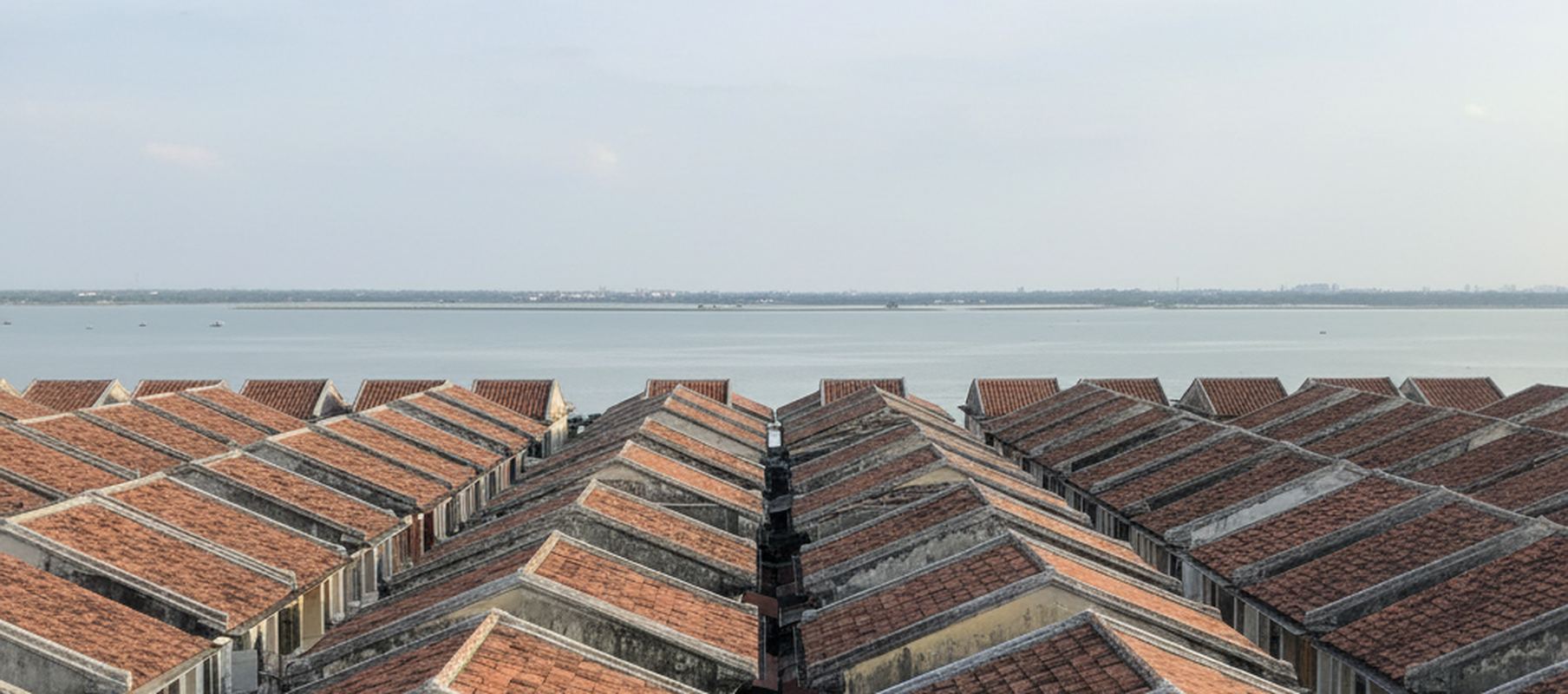Wide rooftop view across rows of two story shophouses with weathered clay tile roofs, the channel water flat beyond them
