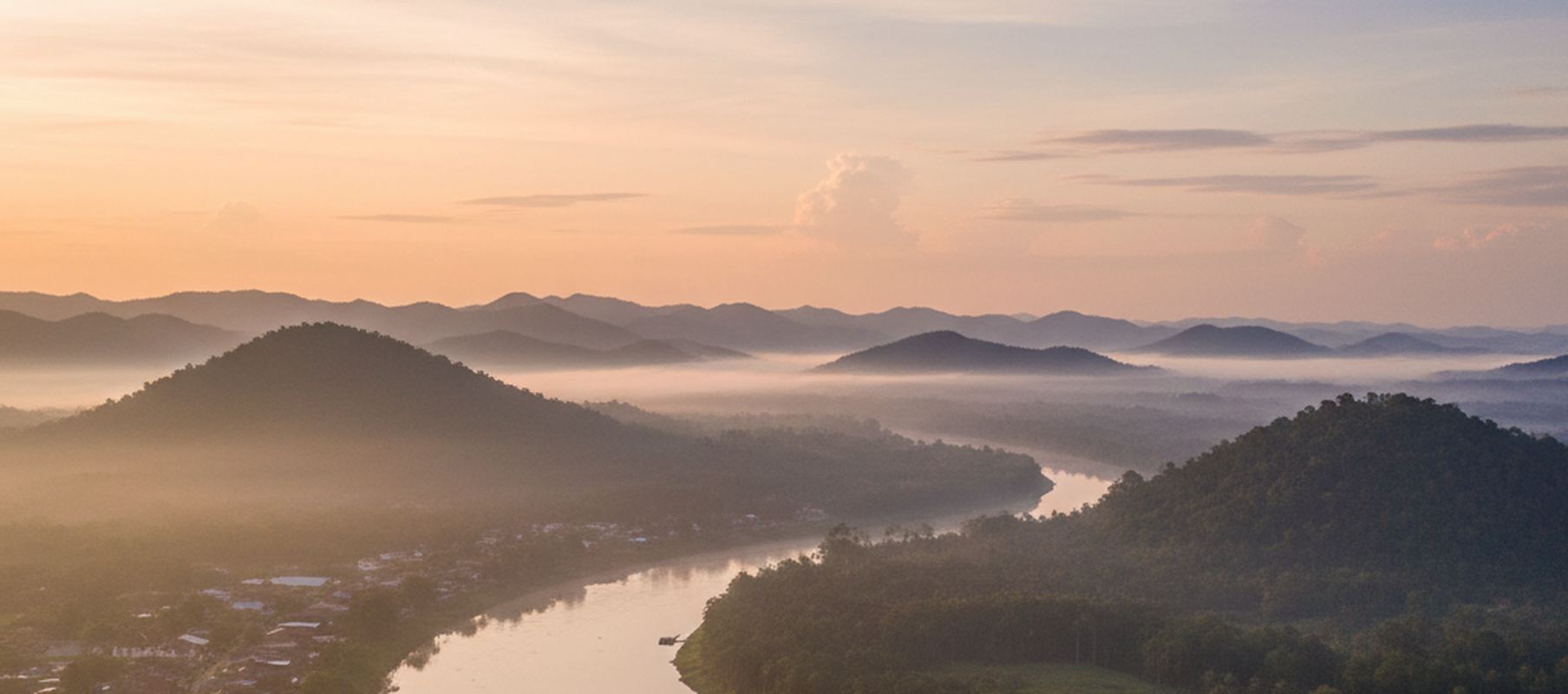 Wide view over the river bend below Kuala Lipis at first light, low town roofs on the near bank and forested ridges running back behind them.
